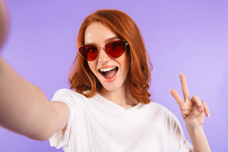 Portrait of a pretty young girl in sunglasses standing isolated over violet background, taking a selfie, showing peaceの写真素材