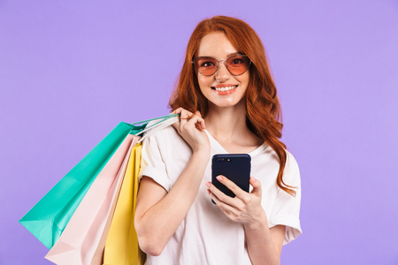 Portrait of a smiling young girl in sunglasses standing isolated over violet background, carrying shopping bags, using mobile phoneの写真素材