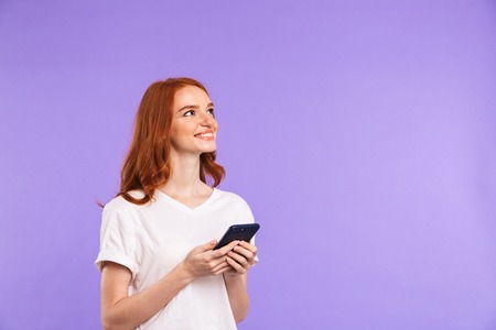 Portrait of a smiling young girl standing isolated over violet background, using mobile phone,...