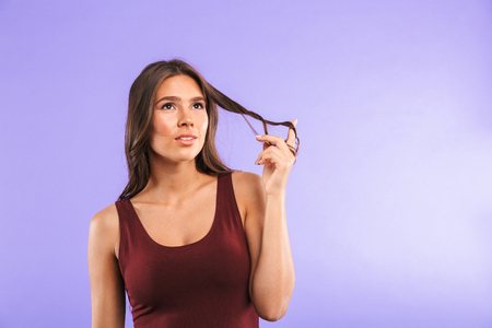 Portrait of a lovely young girl standing isolated over violet background, looking away at copy space, playing with hairの写真素材