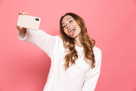 Portrait of a happy young woman taking selfie with mobile phone isolated over pink background, sticking her tongue outの写真素材