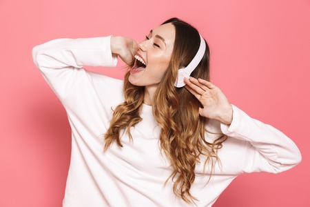 Portrait of a joyful young woman in headphones listening to music isolated over pink background, dancingの写真素材