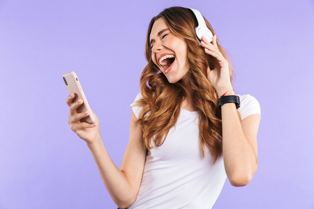 Portrait of a joyful young girl holding mobile phone isolated over violet background, listening to music with headphonesの写真素材