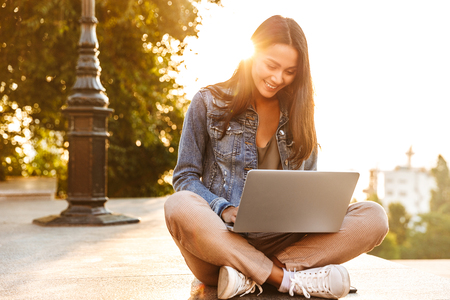 Smiling young asian girl using laptop while sitting on a city streetの写真素材