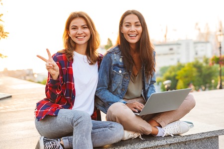 Two happy young girls friends sitting together at the city street, using laptop, showing peaceの写真素材