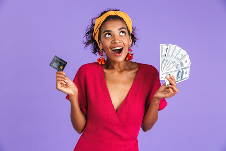Cheerful african woman in dress holding money and credit card while looking up over purple backgroundの写真素材
