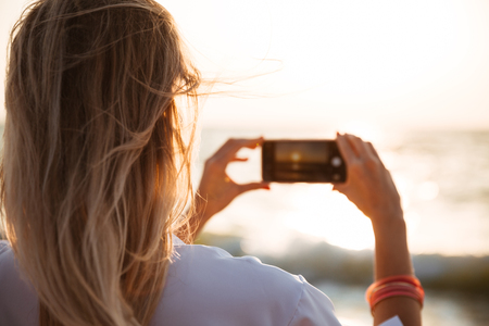 Back view of a woman taking a picture of a sunset at the beach with mobile phoneの写真素材
