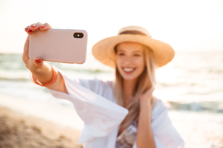 Delighted young girl in summer hat and swimwear spending time at the beach, taking a selfie with outsretched handの写真素材