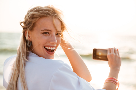 Photo of happy blonde woman 20s smiling and taking photo of sunrise above sea while walking on summer beachの写真素材