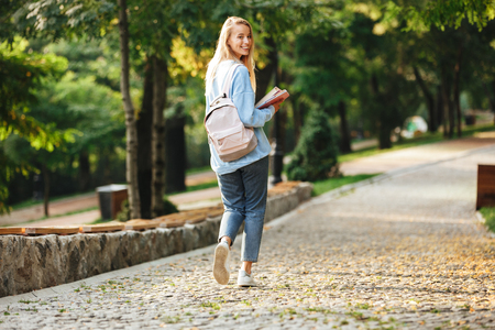 Portrait of a pretty young girl student with backpack and textbook walking at the parkの写真素材