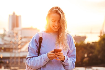 Portrait of a pretty young girl with backpack standing outdoors during sunset, holding mobile phone
