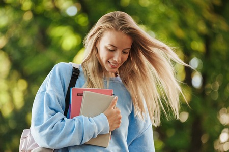 Close up of smiling young girl student with backpack carrying books, walking at the parkの写真素材
