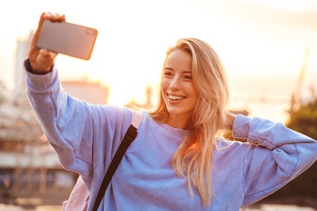 Portrait of a pretty young girl with backpack standing outdoors during sunset, taking selfie with mobile phoneの写真素材