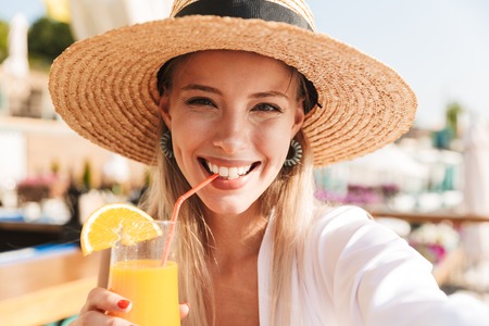 Delighted young girl in summer hat and swimwear resting at the sunny beach, sitting at a cafe with a cocktail, taking a selfieの写真素材