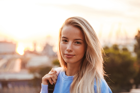 Portrait of a lovely young girl with backpack standing outdoors during sunsetの写真素材