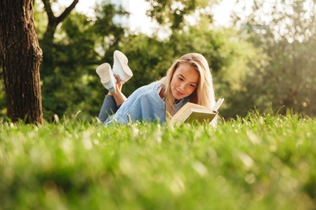 Portrait of a cute young girl laying on a grass at the park, reading a book, taking notesの写真素材