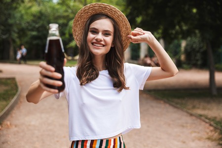 Happy young girl holding bottle with fizzy drink while standing at the park outdoorsの写真素材