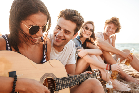 Photo of happy friends loving couple outdoors on the beach sitting while play on the guitar.の写真素材