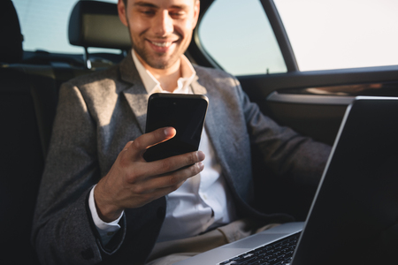 Smiling businessman holding mobile phone while sitting with laptop computer at the back of a carの写真素材
