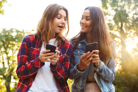 Image of young amazing surprised women friends students walking in the park using mobile phone.の写真素材