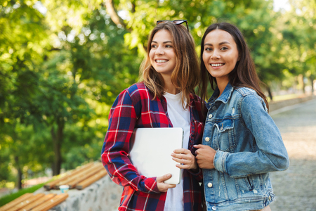 Picture of young amazing ladies students walking in the park holding laptop computer.の写真素材
