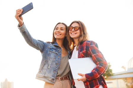 Image of happy young beautiful ladies students walking in the park take a selfie by mobile phone.の写真素材