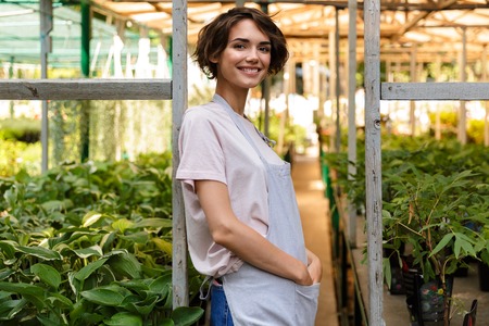 Image of attractive cute woman gardener standing over flowers plants in greenhouse