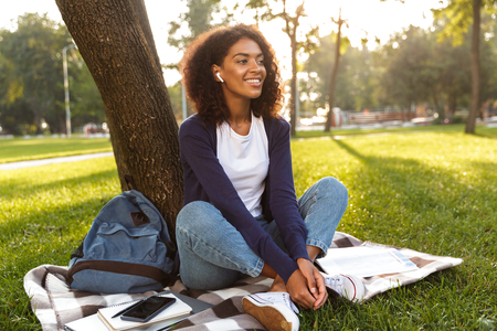 Image of happy african young woman sitting outdoors in park listening music with earphones.の写真素材