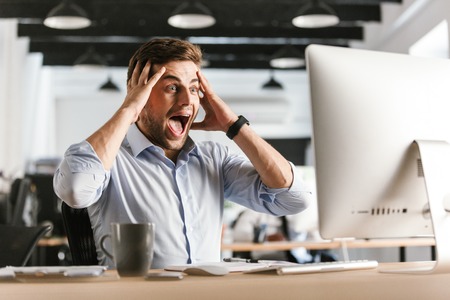 Surprised screaming business man using computer and holding his head while sitting by the table in officeの写真素材