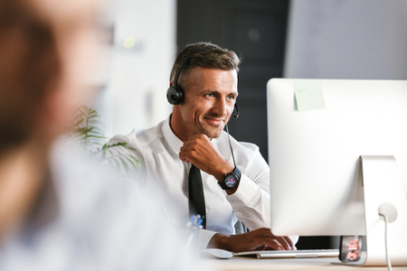 Photo of handsome man 30s wearing office clothes and headset working on computer in call centerの写真素材