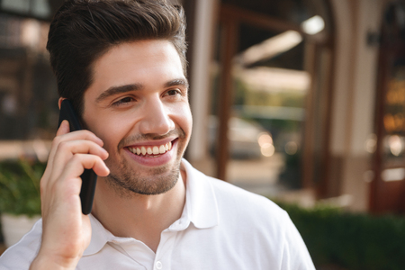 Image of young cheerful man sitting in cafe outdoors while talking by mobile phone.の写真素材