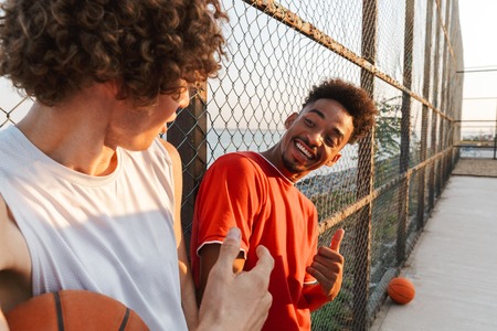 Two young smiling multiethnic men basketball players standing at the sport ground, talkingの写真素材