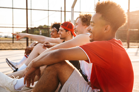 Photo of sporty athletic guys sitting at basketball playground outdoor and watching game during summer sunny dayの写真素材
