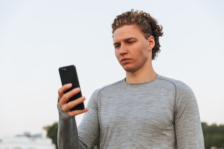 Image of Serious Curly Sportsman using smartphone while being on beach near the seaの写真素材