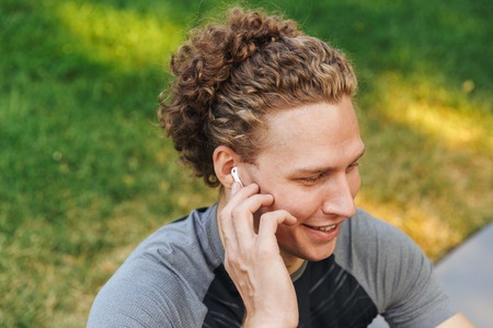 Smiling young sportsman listening to music with earphones while sitting on a fitness mat outdoors