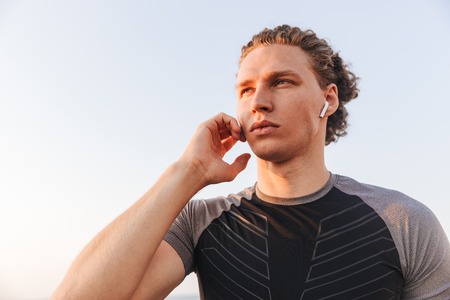 Confident young sportsman in wireless earphones standing outdoors over blue skyの写真素材