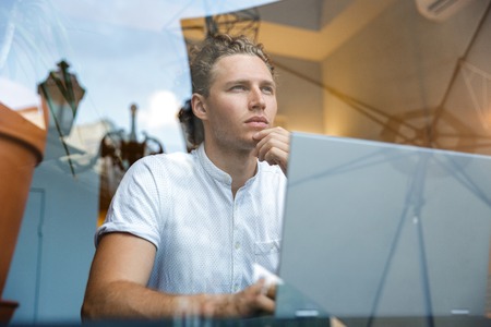 Pensive curly business man looking away while sitting by the table with laptop computer in cafe