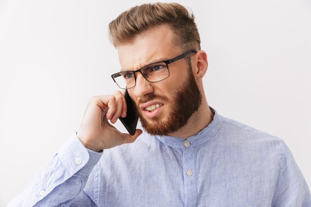 Portrait of an angry young bearded man in eyeglasses standing isolated over white, talking on mobile...