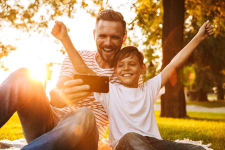 Image of happy young man father have a rest with his son outdoors in park play games with mobile phone.の写真素材