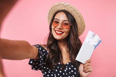 Cheerful brunette woman in straw hat and sunglasses making selfie while holding passport with tickets over pink backgroundの写真素材