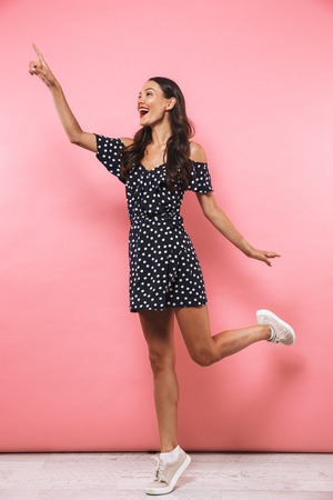 Full length image of Pleased brunette woman in dress jumping while pointing and looking away over pink background