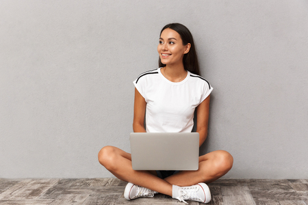 Image of happy young woman wearing casual clothing using silver laptop, while sitting on floor with legs crossed isolated over gray backgroundの写真素材
