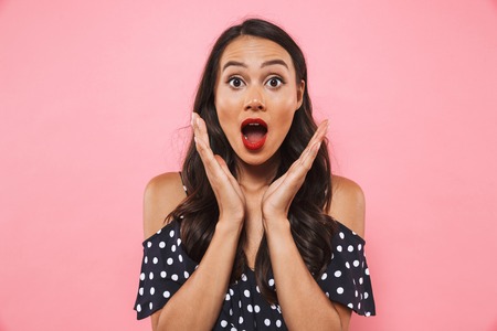 Surprised brunette woman in dress looking at the camera with open mouth over pink backgroundの写真素材