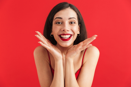 Portrait of an excited young woman in dress isolated over red background, holding hands at her faceの写真素材