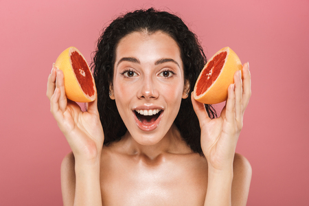 Beauty photo of european brunette woman with long hair smiling and holding pieces of grapefruit isolated over pink backgroundの写真素材