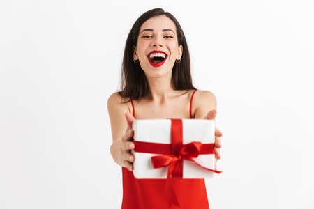 Portrait of a joyful young woman in red dress holding present box isolated over white background