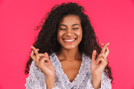 Portrait of a smiling young african woman in summer dress standing isolated over pink background, holding fingers crossedの写真素材