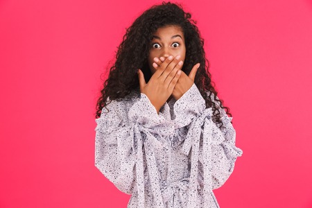 Portrait of a shocked young african woman in summer dress standing isolated over pink background, cover mouthの写真素材