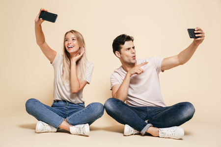 Photo of cute happy young loving couple sitting isolated over beige wall background make selfie by mobile phones.の写真素材