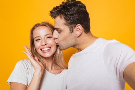 Photo of happy excited young loving couple standing isolated over yellow wall background take a selfie by camera kissing.の写真素材